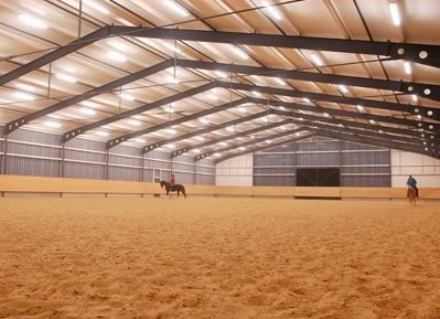 Blick in die Reithalle der Reitsportanlage Haldau — zwei Reihen Deckenlichter über dem Sandboden, im Hintergrund Reiter